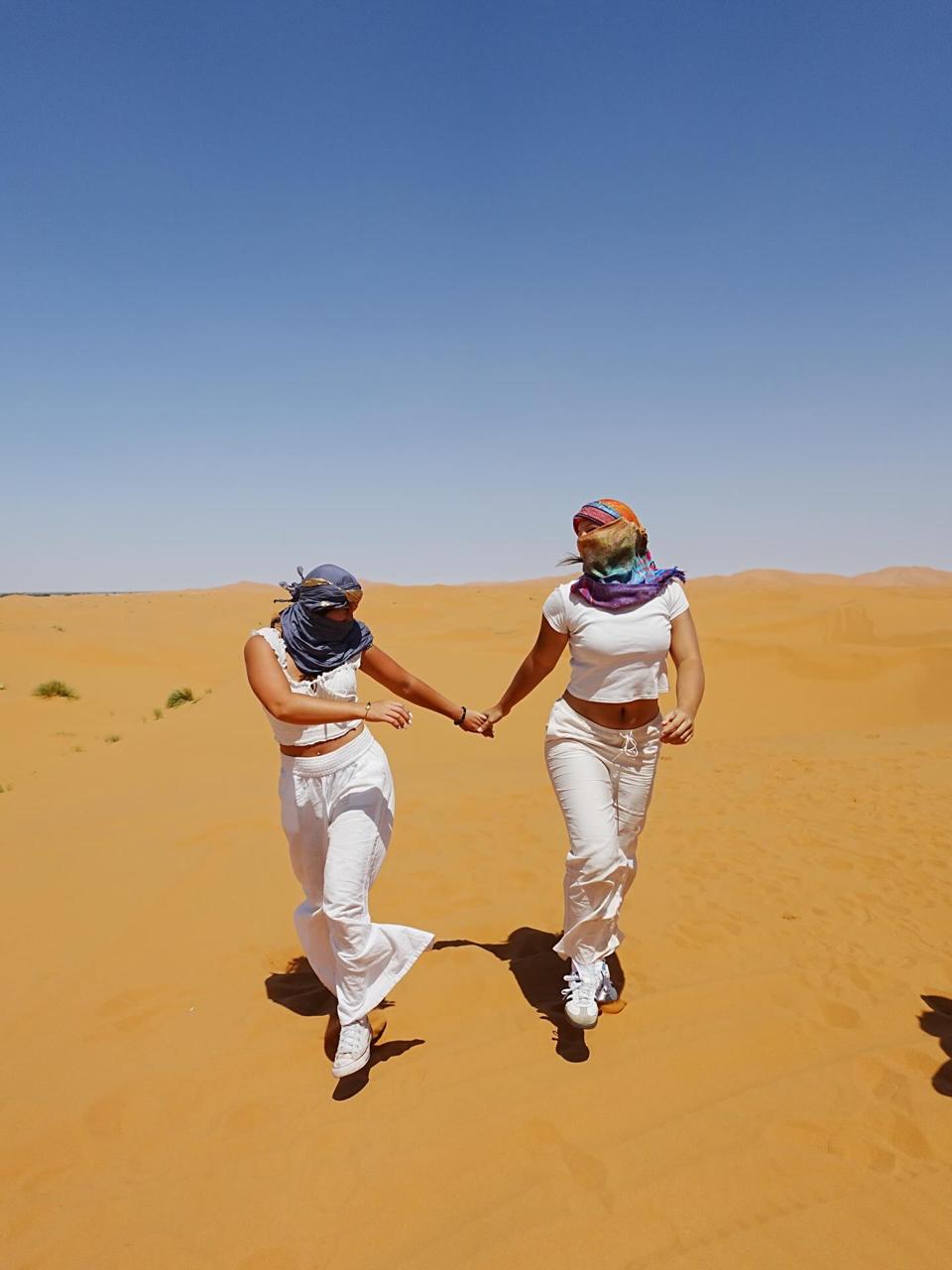 Two friends in white outfits and scarves holding hands and running across the sand