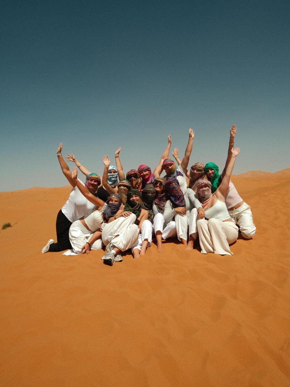 Large group sitting together on a dune with their arms raised, wearing colorful scarves