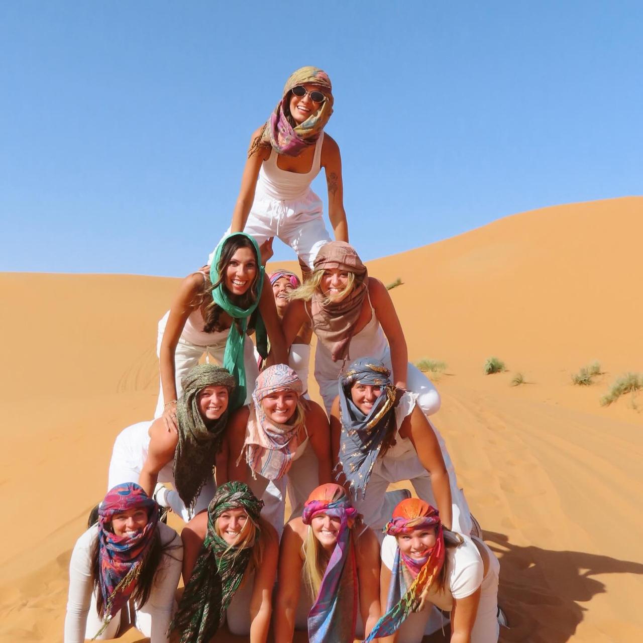 Group of students in colorful scarves stacked in a human pyramid on an orange Sahara dune