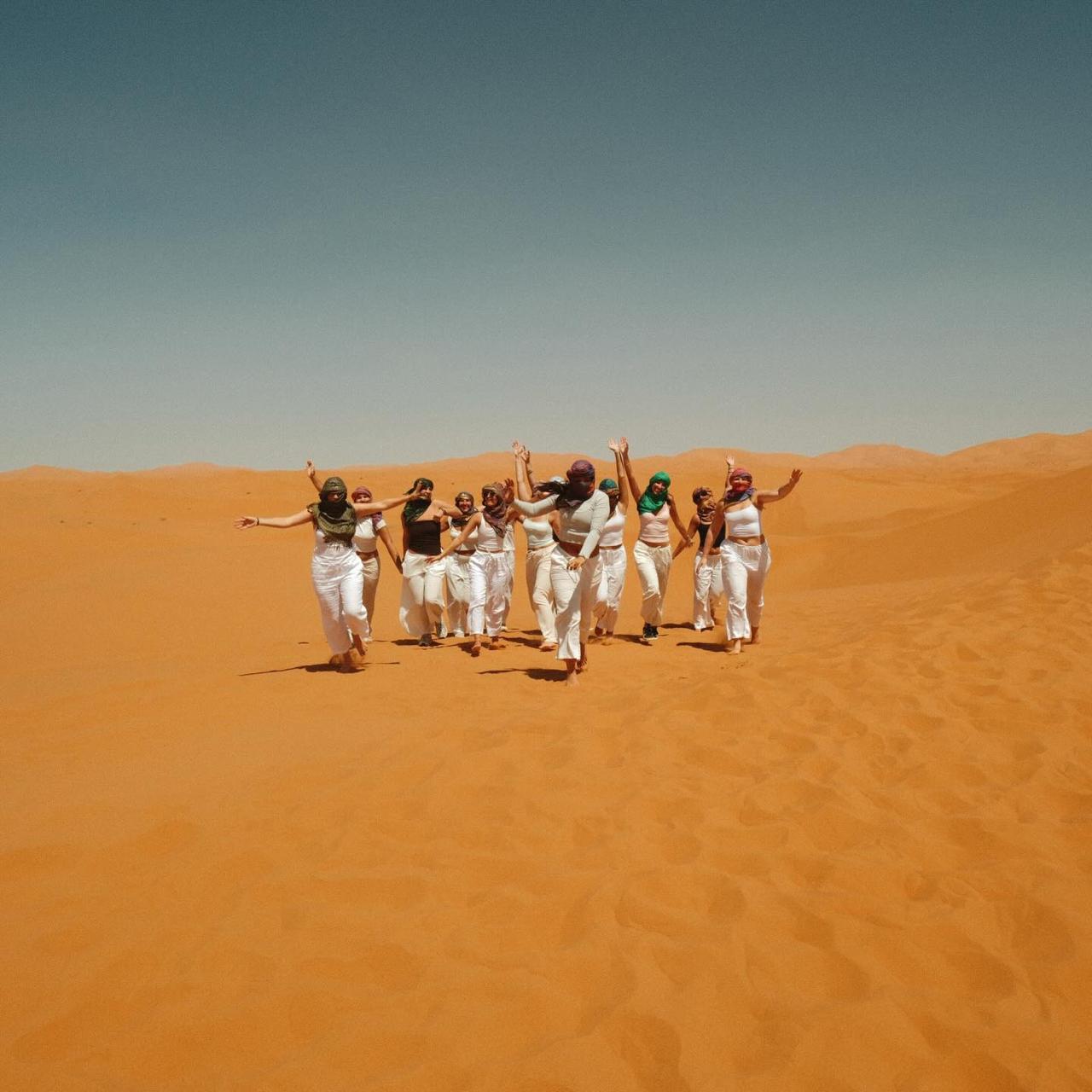 Group of students in white outfits and scarves running and cheering across an open Sahara dune
