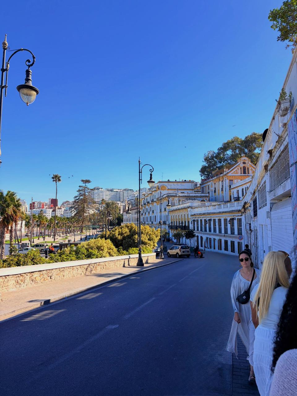 Two women walking on a sunny street lined with white buildings and palm trees