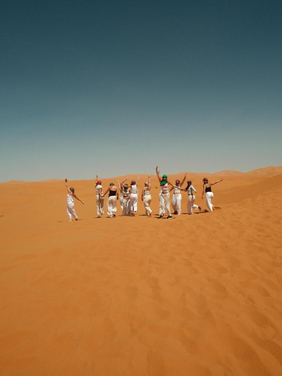 Distant group of students jumping and posing on a vast orange dune under a clear sky