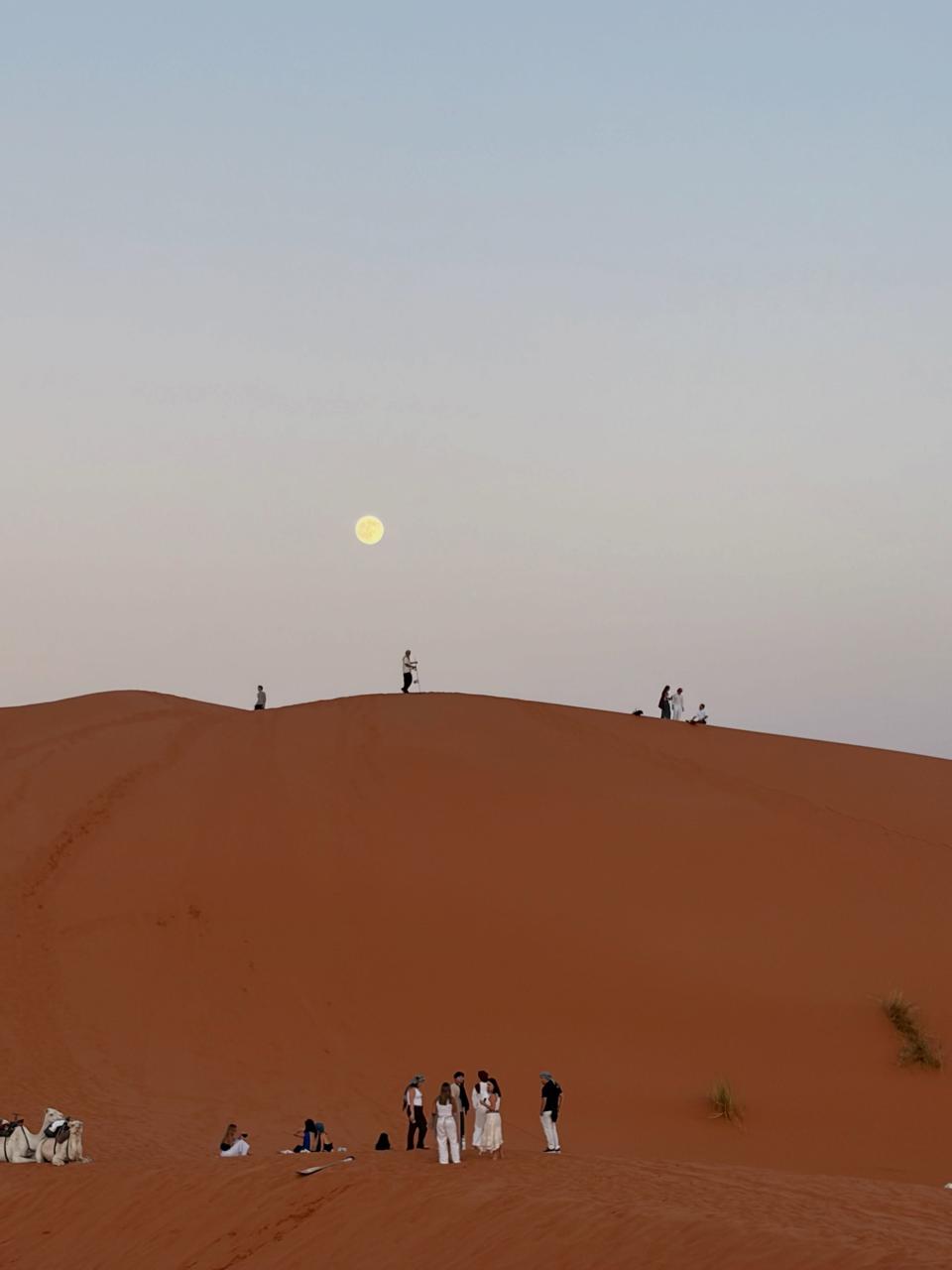 Small group of people on a dune ridge at dusk with a pale moon rising